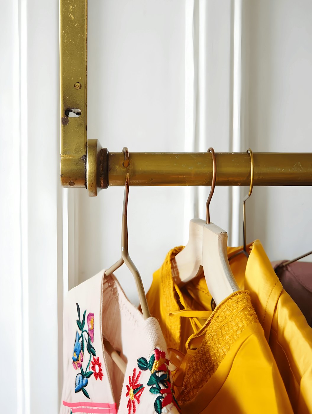 Embroidered vintage blouses and a mustard yellow jacket hanging from a brass clothing rail against a white panelled wall